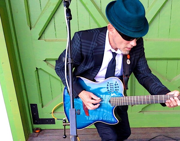 Man in suit with hat and sunglasses playing a blue guitar in front of a green door.